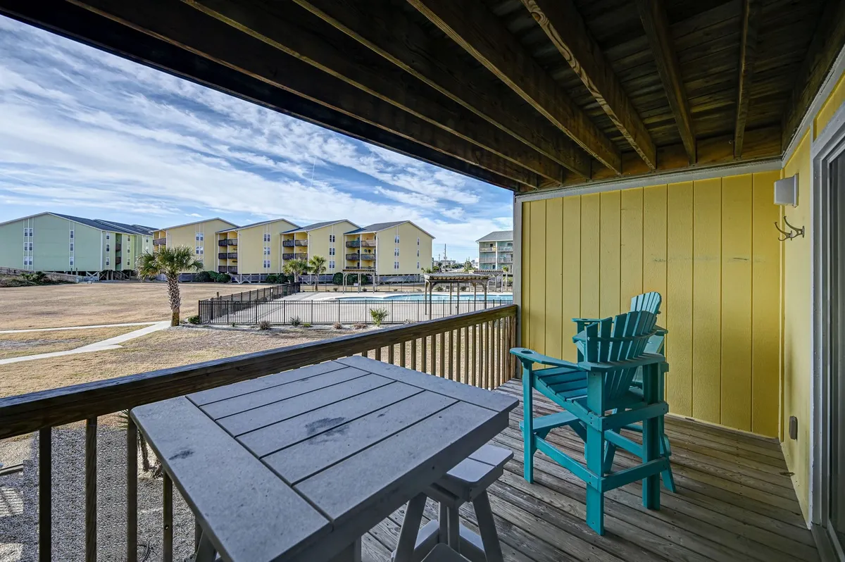 An exterior shot from the perspective of a private porch or balcony, featuring a turquoise Adirondack chair and wooden table. The swimming pool and vibrant yellow and green resort buildings are visible in the distance, capturing a relaxed vantage point for enjoying a drink or watching pool activities.