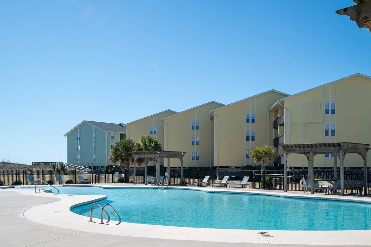 This vibrant exterior photo features a sparkling swimming pool surrounded by colorful resort buildings, palm trees, inviting lounge chairs, and shaded pergolas. The scene is lively yet relaxing, capturing guests sunbathing, swimming, and socializing under sunny skies.