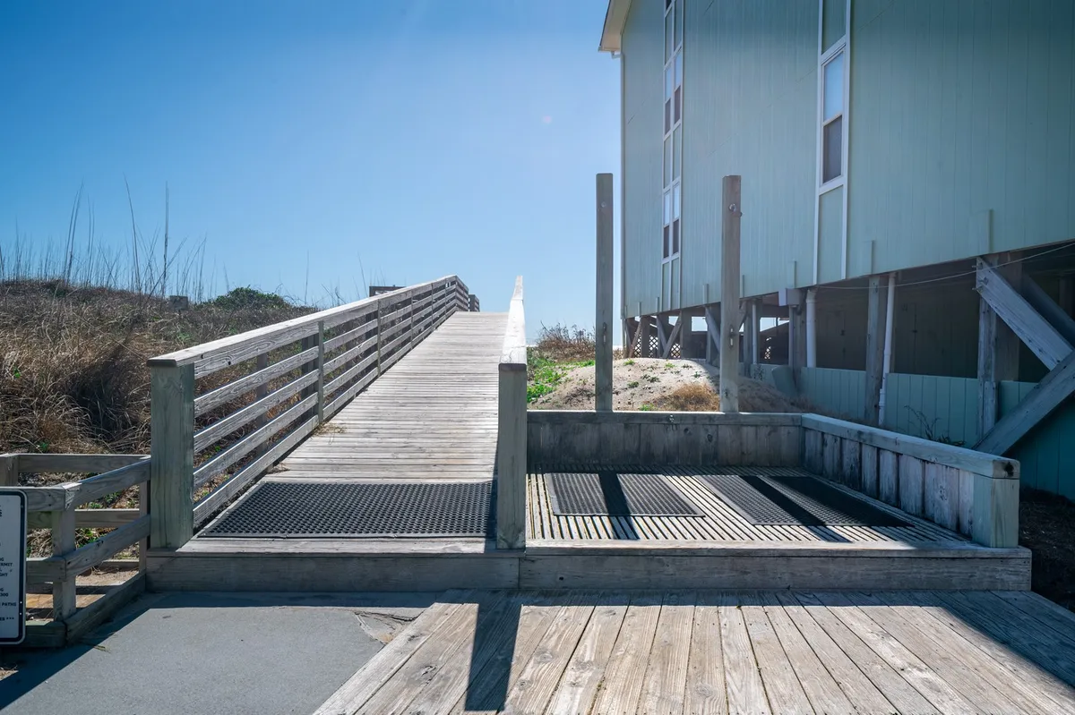 A wooden ramp leads through lush coastal vegetation directly toward the sandy shore. The 'beach access' sign marks the way, creating an inviting pathway for guests to begin their beach adventure. The scene feels tranquil and welcoming, emphasizing proximity and ease of access.
