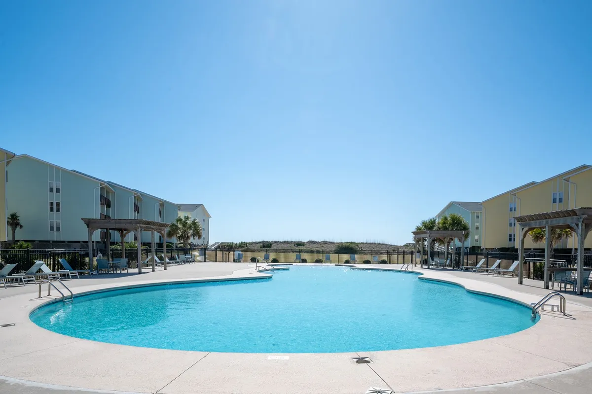 A tranquil swimming pool scene featuring lounge chairs, surrounding buildings, and a bright, clear sky. The calm water reflects the late afternoon sunlight, inviting guests to unwind and enjoy peaceful moments outdoors.