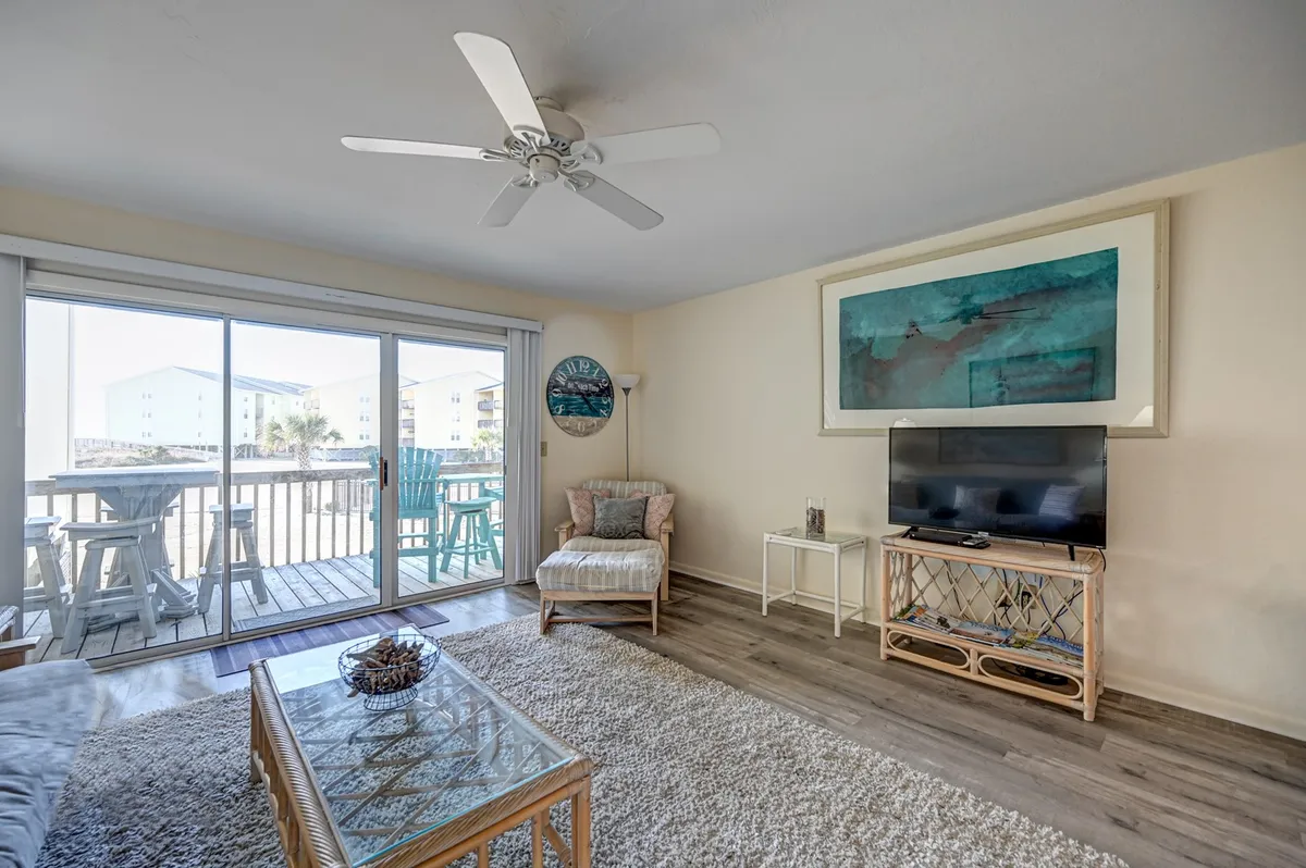 A bright living area with a large sliding glass door lets natural light flood in. The space features a comfortable chair, coffee table, and a television, with artwork adding color and character. The sliding glass door opens to a balcony, visually connecting the interior with the outdoors.