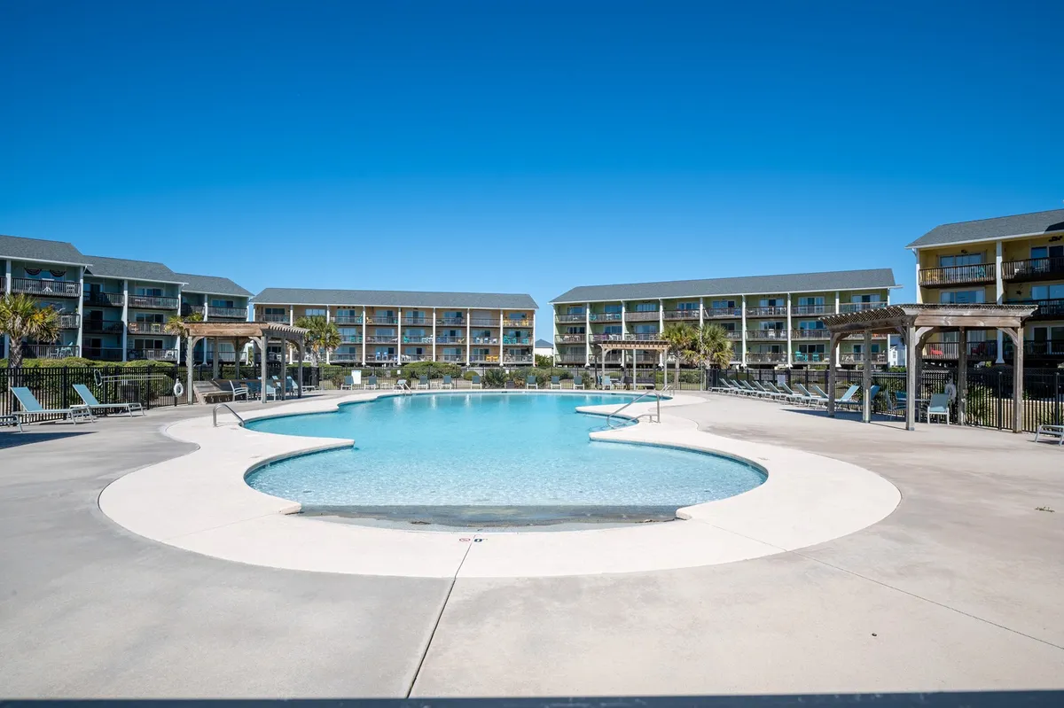 A sparkling swimming pool surrounded by lounge chairs, palm trees, and colorful buildings. The area is bright, inviting, and designed for both relaxation and socializing, capturing the essence of a community pool in a resort-like setting.
