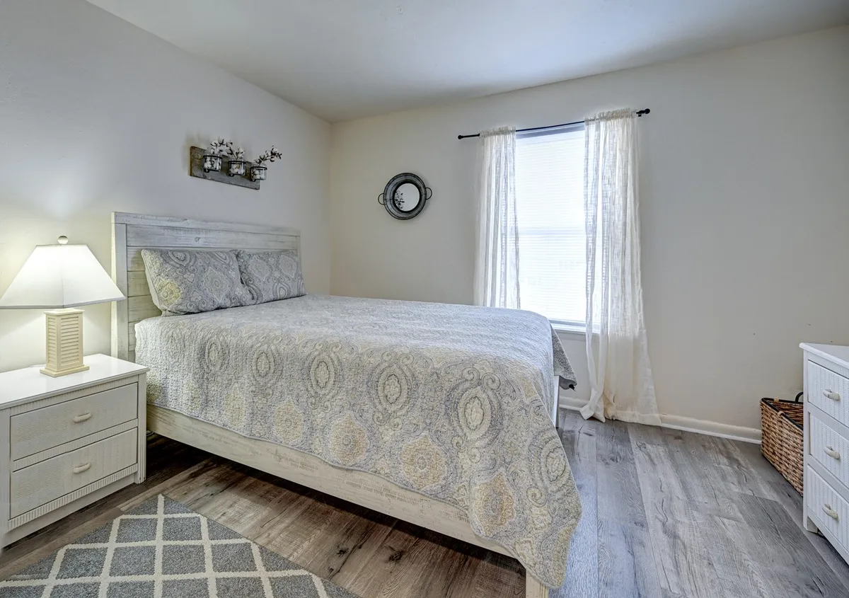A serene queen bedroom with patterned bedding, a nightstand and lamp for evening reading, and a window draped in sheer curtains that lets in gentle natural light. The space feels inviting and peaceful, perfect for relaxation and privacy.