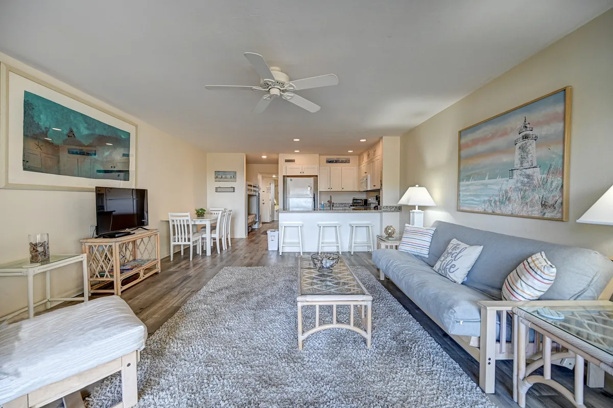 A light-filled living room with a comfortable, light-colored sofa (which doubles as a pull-out sleeper), a central coffee table, wall art, and an adjacent dining area. The setting feels inviting for relaxation or communal gatherings.