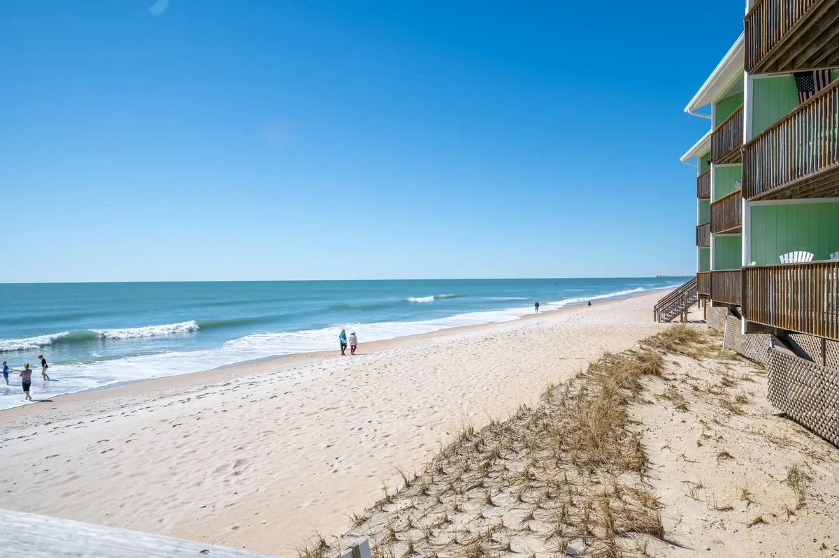 A tranquil beach scene with gentle ocean waves, people walking along the shore, and a nearby green building. The setting captures joyful family walks, sunbathing, and the quintessential beach experience, with the condo complex visible in the background.