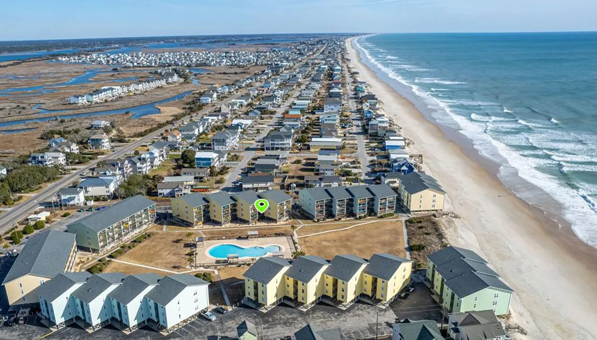 A wide exterior view showcasing the beachfront, swimming pool, a row of resort houses, and rolling ocean waves in the background. The scene evokes family gatherings by the pool and easy transitions between the beach, pool, and living space.