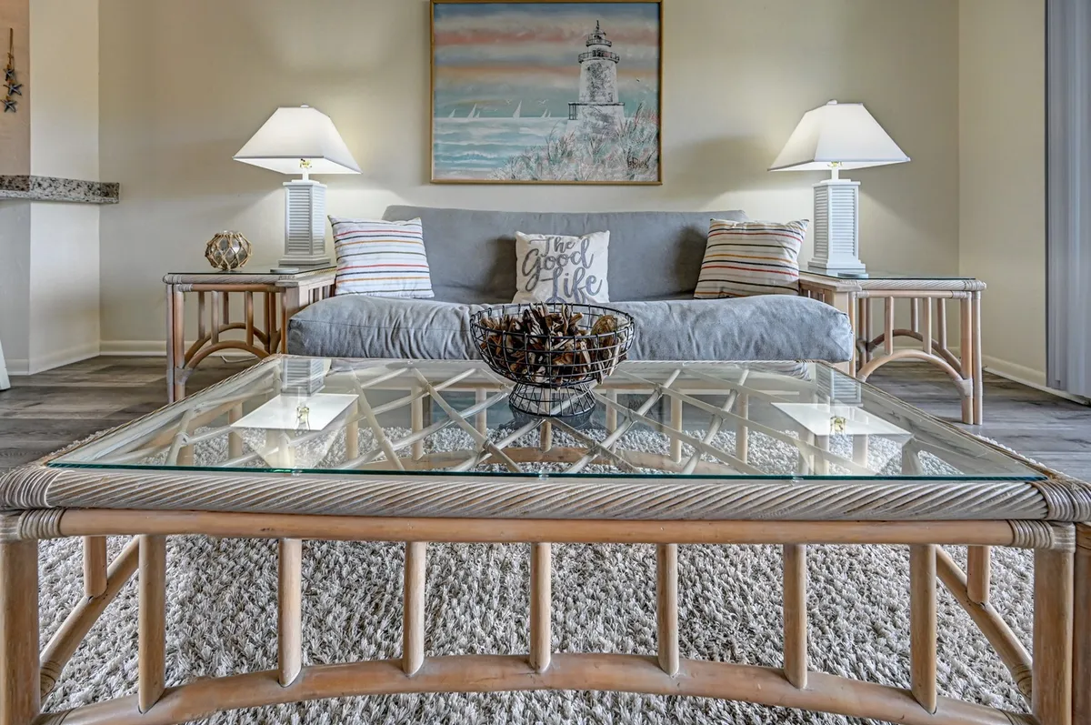A sunlit, inviting living room featuring a plush sofa, elegant coffee table, coastal-inspired artwork, and soft ambient lighting from stylish lamps. The cozy setup offers a perfect gathering spot for relaxing with loved ones or unwinding after a day at the beach.
