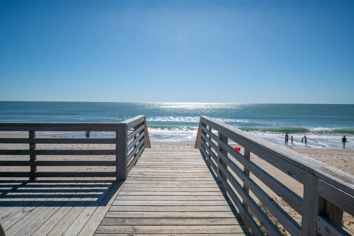 A wooden walkway extends out over the dunes to the beach, with the ocean and distant beachgoers visible in the background. The scene invites exploration and relaxation, illustrating both beach access and the social atmosphere.