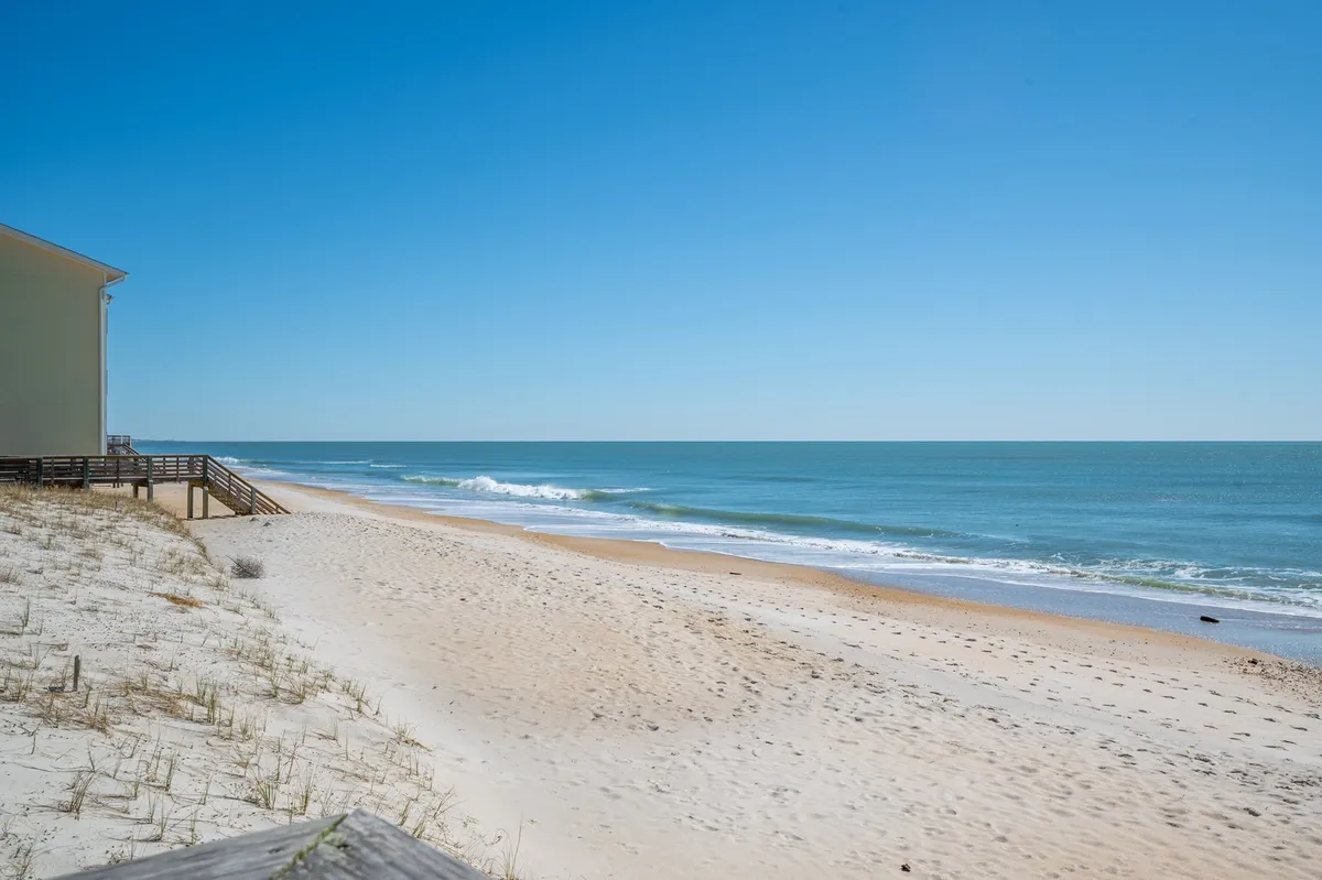 An expansive view of the beach shows soft sand, rolling ocean waves, and a wide blue sky. The boardwalk is visible, hinting at both classic beach pastimes and the walkable connection to local attractions. The scene is serene, vibrant, and inviting.